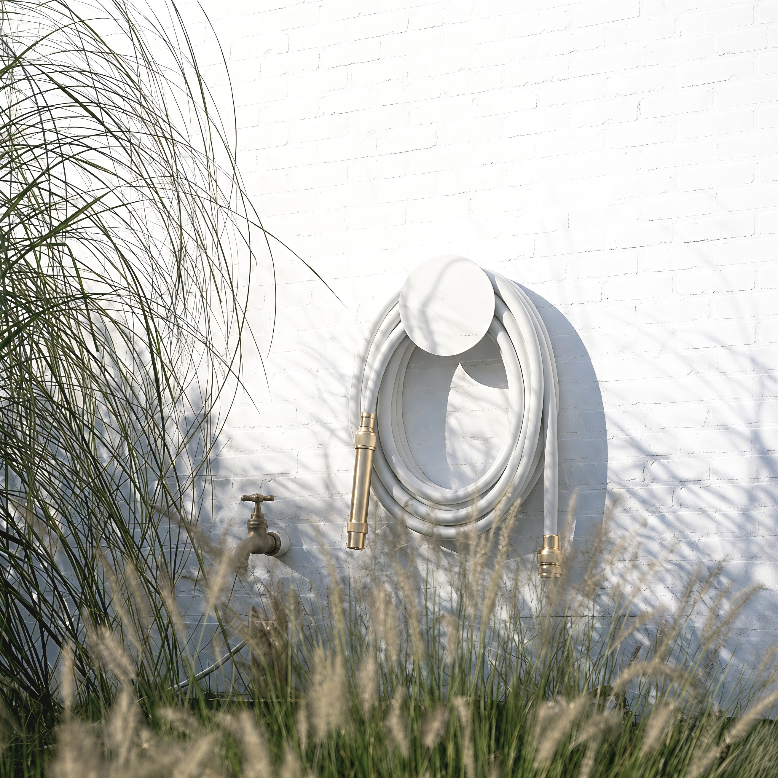 Large white hose holder with pale grey garden hose mounted on a white brick wall among ornamental grasses.