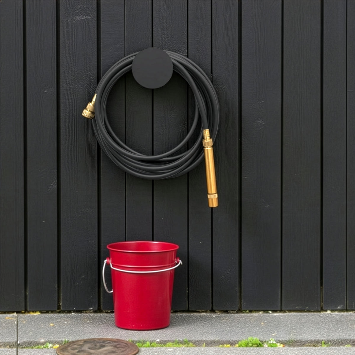 Small black hose holder with coiled black garden hose mounted on a charcoal timber fence above a red bucket.