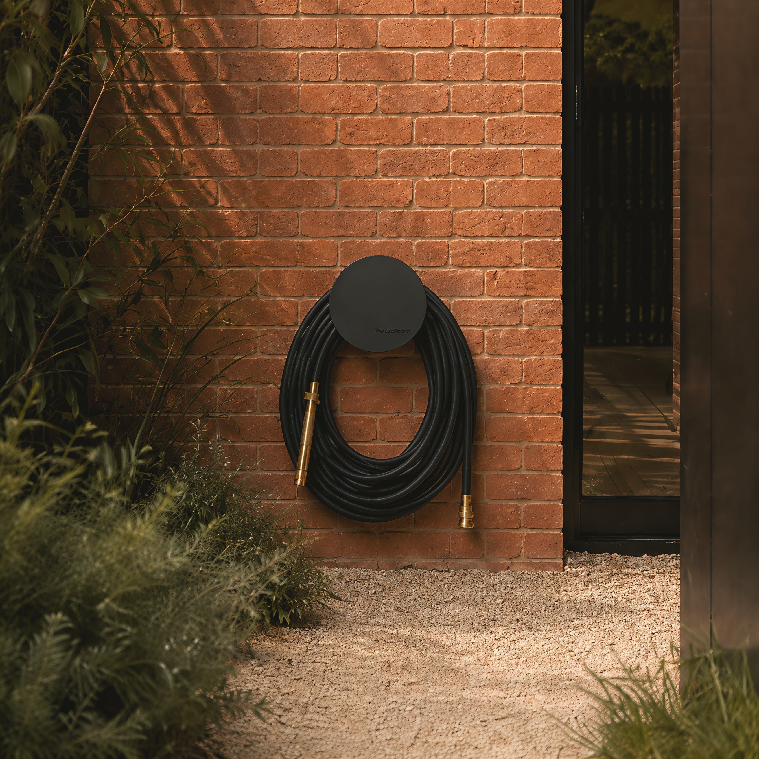 Matte black hybrid garden hose mounted on a black hose holder against a warm brick garden wall.