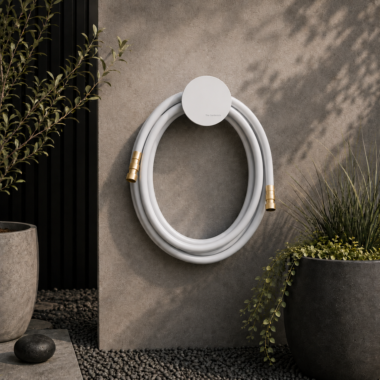 Modern outdoor garden scene featuring a light grey premium garden hose with solid brass fittings mounted on a minimalist white circular hose holder with subtle The Gardenista branding, set against a textured architectural wall with soft natural sunlight, dark stone planters, olive tree foliage, and black pebble landscaping.